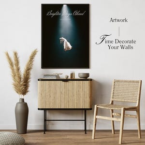 Puede incluir: Un póster enmarcado con el texto "Brighter Days Ahead" y una foto de una mujer con un vestido blanco flotando en el aire. El póster está colgado en una pared blanca sobre un armario de color marrón claro con un marco negro. Una silla de mimbre está en primer plano.