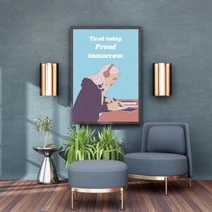 Op de afbeelding: Een ingelijste print met de tekst "Tired today. Proud tomorrow." hangt boven een blauwe stoel en poef. De print toont een illustratie van een persoon met een koptelefoon die schrijft. De kamer heeft een modern design met gouden accenten en planten.