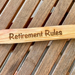 May include: A wooden sign with the words "Retirement Rules" engraved on it. The sign is painted pink at one end, resembling a pencil.
