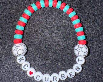 May include: A red and turquoise beaded bracelet with white letter beads spelling "KC CURRENT" and a heart bead. The bracelet also has two white football beads.