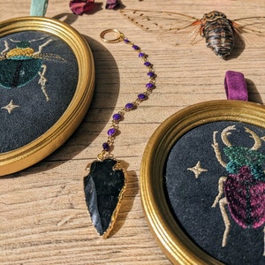May include: Two circular embroidery hoops with black fabric and gold frames. The hoops feature embroidered images of insects. A gold chain with purple beads hangs from the top hoop and has a black arrowhead pendant.