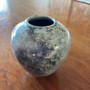 May include: A small, round, silver-toned vase with a dark interior. The vase has a weathered appearance with dark spots and a slightly textured surface. The vase sits on a wooden surface.