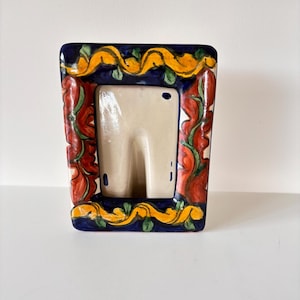 May include: A rectangular ceramic picture frame with a cream-colored interior. The frame is decorated with a blue border, red floral accents, and yellow and green decorative elements. The frame is designed to hold a small photograph or artwork.