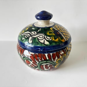 May include: A ceramic jar with a lid, featuring a vibrant hand-painted design. The jar's base showcases a pattern of red, green, and white floral motifs. The lid is adorned with blue, green, yellow, and white patterns. The jar has a blue knob on top.