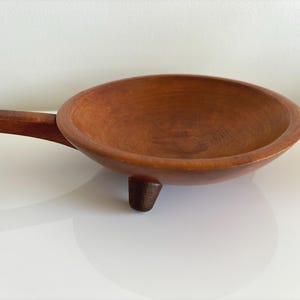 May include: A wooden bowl with a long handle and a small, triangular foot. The bowl and handle are a warm brown color, and the bowl has a rounded shape. The bowl is likely used for serving or cooking.
