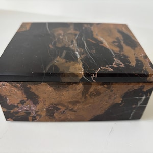 May include: A square jewellery box with a hinged lid. The box is made of black marble with brown and white veining. The lid is flat and polished, and the sides are angled.