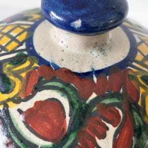 May include: Close-up of a ceramic lid with a blue knob. The lid features a colorful, hand-painted design with red, green, yellow, and blue patterns. The design includes floral and geometric elements, creating a vibrant and artistic appearance. The lid is likely for a decorative or functional ceramic piece.