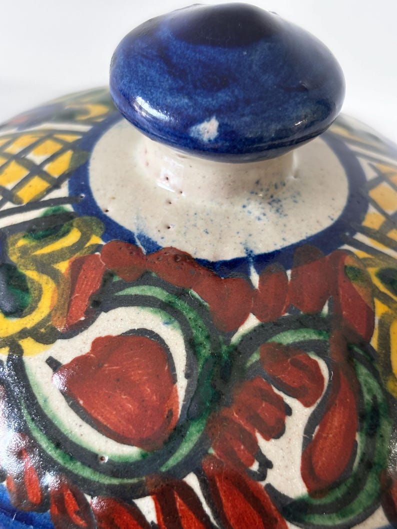 May include: Close-up of a ceramic lid with a blue knob. The lid features a colorful, hand-painted design with red, green, yellow, and blue patterns. The design includes floral and geometric elements, creating a vibrant and artistic appearance. The lid is likely for a decorative or functional ceramic piece.