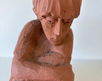 May include: A terracotta-colored sculpture of a person in a seated position, with arms crossed and head bowed. The figure has a textured, carved appearance, with visible brushstrokes and a matte finish. The sculpture is a representation of human form.