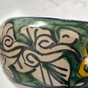 May include: Close-up of a ceramic bowl with a hand-painted floral design. The bowl features a cream-colored base with green and black leaf patterns. The rim is painted with a dark blue and green border, and yellow and blue flowers are visible.