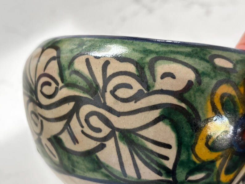 May include: Close-up of a ceramic bowl with a hand-painted floral design. The bowl features a cream-colored base with green and black leaf patterns. The rim is painted with a dark blue and green border, and yellow and blue flowers are visible.