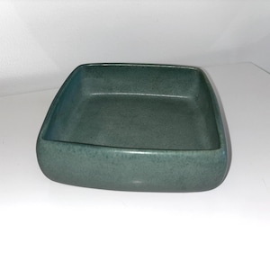 May include: A square, teal ceramic dish with rounded corners. The dish has a speckled texture and a smooth interior. The sides are slightly sloped, and the overall design is simple and modern. Suitable for serving food or as a decorative item.