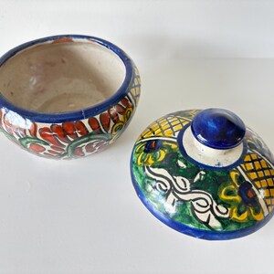 May include: Open ceramic bowl and lid with a blue rim and knob. The bowl features a floral design with red, green, and yellow accents. The lid has a similar design with a blue knob.