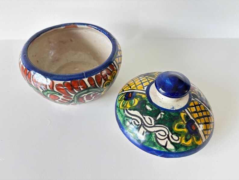May include: Open ceramic bowl and lid with a blue rim and knob. The bowl features a floral design with red, green, and yellow accents. The lid has a similar design with a blue knob.
