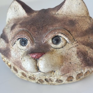 Könnte beinhalten: Eine Keramikfigur in Form eines Katzenkopfes mit gesprenkelter braun-cremefarbener Oberfläche und dunkelbraunen Tabby-Streifen. Die Katze hat markante Augen, eine texturierte rosa Nase und eine dekorative Reihe kleiner, kreisförmiger Vertiefungen.