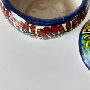 May include: A partially visible, hand-painted ceramic bowl with a lid. The bowl's rim is dark blue with red and green floral designs. The lid features a yellow and blue pattern with a white and green design. The bowl is on a white surface.