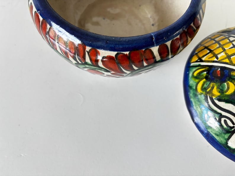 May include: A partially visible, hand-painted ceramic bowl with a lid. The bowl's rim is dark blue with red and green floral designs. The lid features a yellow and blue pattern with a white and green design. The bowl is on a white surface.