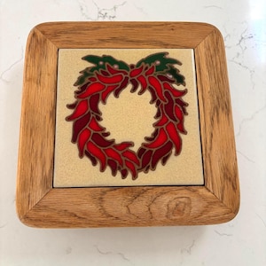 Puede incluir: Un salvamanteles cuadrado con un marco de madera clara que rodea un azulejo de cerámica central. El azulejo muestra una ilustración de una corona decorativa compuesta por chiles rojos y hojas verdes sobre un fondo crema.