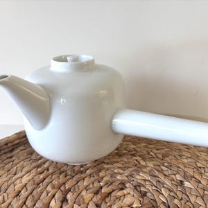 May include: A white ceramic teapot with a cylindrical handle and a spout. The teapot is sitting on a woven, brown, circular placemat. The teapot has a rounded body and a small lid.