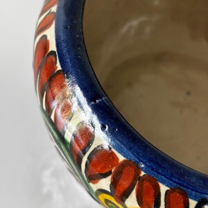 May include: Close-up of a ceramic bowl with a vibrant floral design. The rim is painted with a deep blue band, and the exterior features red, yellow, and green floral patterns against a cream background. The interior is a light beige color.