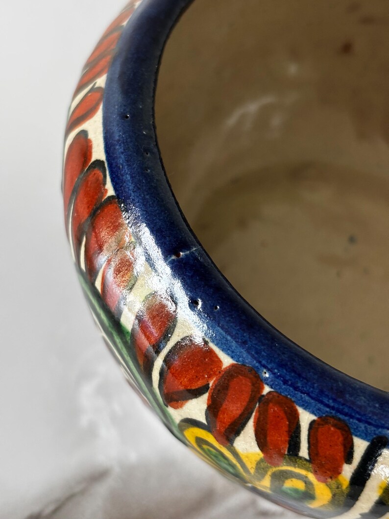 May include: Close-up of a ceramic bowl with a vibrant floral design. The rim is painted with a deep blue band, and the exterior features red, yellow, and green floral patterns against a cream background. The interior is a light beige color.