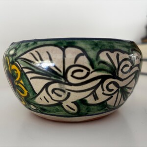 May include: A small, hand-painted ceramic bowl with a floral design. The bowl features a cream-colored base with green, yellow, and blue accents. The rim is dark green, and the bowl has a rounded shape.