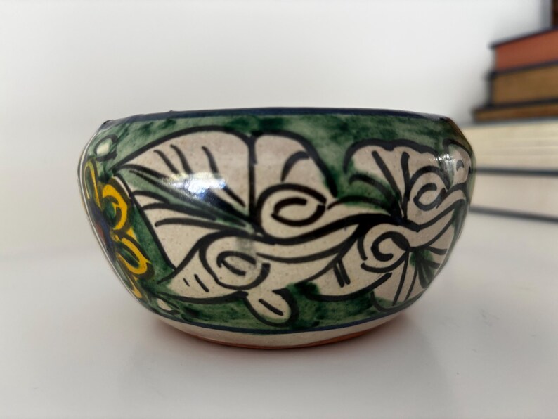 May include: A small, hand-painted ceramic bowl with a floral design. The bowl features a cream-colored base with green, yellow, and blue accents. The rim is dark green, and the bowl has a rounded shape.