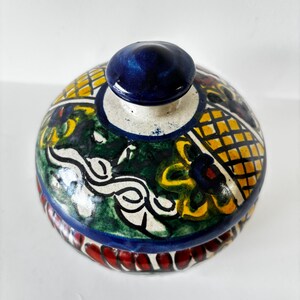 May include: A hand-painted ceramic jar with a lid. The jar features a vibrant design with yellow, green, and blue patterns. The lid has a dark blue knob. The base of the jar has a red and white design.