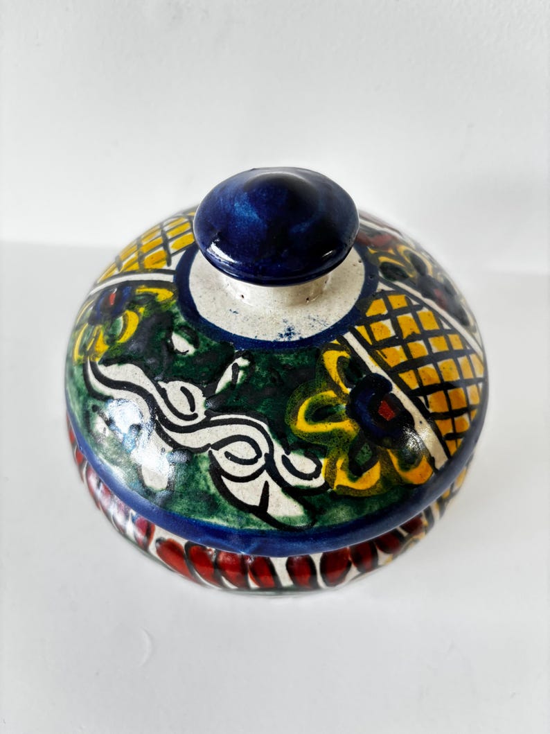 May include: A hand-painted ceramic jar with a lid. The jar features a vibrant design with yellow, green, and blue patterns. The lid has a dark blue knob. The base of the jar has a red and white design.