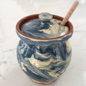 May include: A ceramic honey pot featuring a marbled blue and cream glaze with a smooth, glossy finish. The pot includes a matching lid with a central knob and a small opening for the included light-colored wooden honey dipper stick.