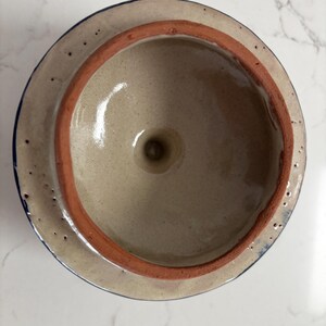 May include: A ceramic lid with a light beige interior and a reddish-brown rim. The lid's exterior is a lighter beige with a dark blue trim. The lid has a small hole in the center.
