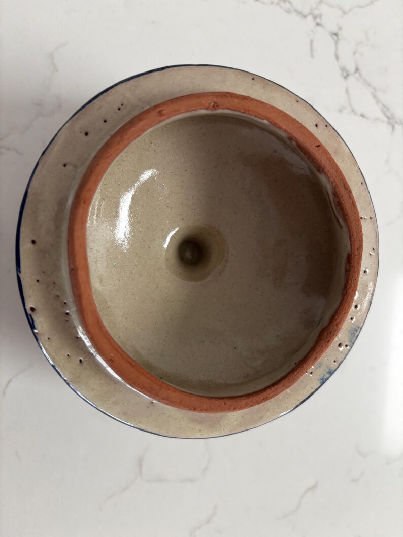 May include: A ceramic lid with a light beige interior and a reddish-brown rim. The lid's exterior is a lighter beige with a dark blue trim. The lid has a small hole in the center.