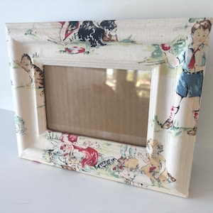 May include: A rectangular picture frame covered in a cream-colored fabric featuring a printed pattern of children playing outdoors, a cat, and green grass. The frame has a beveled edge and a central rectangular opening with a brown cardboard backing.