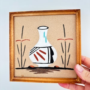 May include: A framed sand art piece depicting a white vase with black and red details. The vase is set against a beige background with plant motifs. The artwork is enclosed in a wooden frame.