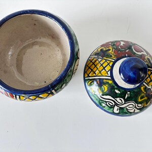 May include: A ceramic bowl with a lid, featuring a vibrant, hand-painted design. The bowl's interior is beige, with a blue rim and colorful exterior patterns. The lid has a blue knob and a decorative pattern with yellow, green, and red accents.