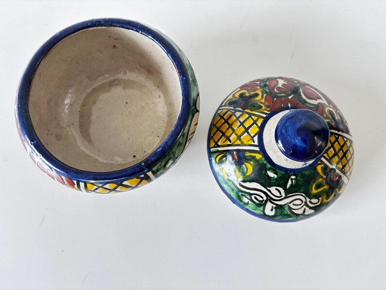 May include: A ceramic bowl with a lid, featuring a vibrant, hand-painted design. The bowl's interior is beige, with a blue rim and colorful exterior patterns. The lid has a blue knob and a decorative pattern with yellow, green, and red accents.