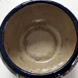 May include: A handmade ceramic bowl with a dark blue rim and a light brown interior. The bowl has a round shape and appears to be glazed. The exterior has decorative elements in red, yellow, and blue.