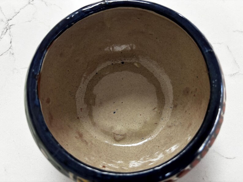 May include: A handmade ceramic bowl with a dark blue rim and a light brown interior. The bowl has a round shape and appears to be glazed. The exterior has decorative elements in red, yellow, and blue.