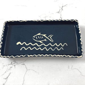 Puede incluir: Una bandeja rectangular de cerámica azul marino con un diseño de pez y olas en blanco. El borde de la bandeja tiene un borde festoneado blanco. Esta bandeja decorativa es adecuada para servir o exhibir.