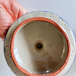 May include: A ceramic lid with a speckled beige interior and a reddish-orange rim. The outer edge is white with blue accents and small, evenly spaced holes. The lid is designed to fit a container.