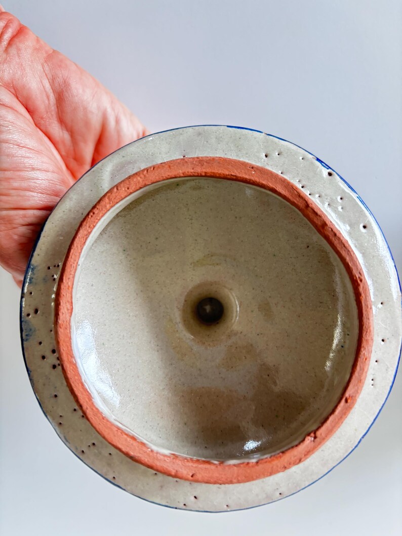 May include: A ceramic lid with a speckled beige interior and a reddish-orange rim. The outer edge is white with blue accents and small, evenly spaced holes. The lid is designed to fit a container.