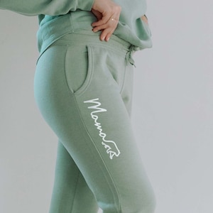 Może przedstawiać: Jasne zielone spodnie dresowe z białym logo "Mama Bear" na lewej nogawce. Spodnie dresowe są podwinięte przy kostkach, a osoba, która je nosi, ma na sobie białe buty sportowe.