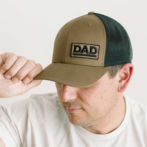 Może przedstawiać: Brązowa i czarna czapka z daszkiem z czarną naszywką z napisem "DAD" z przodu.