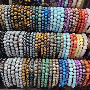 May include: A collection of colourful gemstone bracelets, including tiger's eye, amethyst, turquoise, and jasper. The bracelets are arranged in rows on a wooden surface.