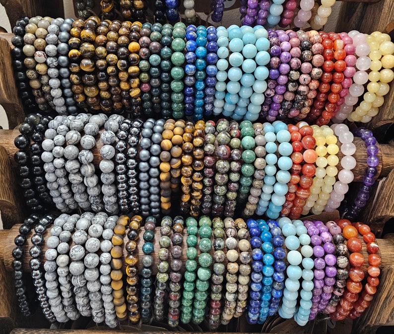 May include: A collection of colourful gemstone bracelets, including tiger's eye, amethyst, turquoise, and jasper. The bracelets are arranged in rows on a wooden surface.