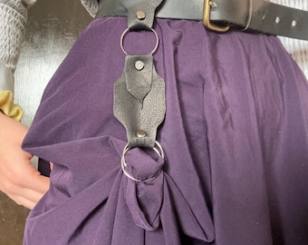 May include: A black leather belt with a silver buckle and a decorative leather strap with a silver ring hanging from it. The belt is worn over a purple skirt.