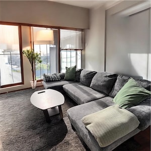 May include: A modern living room with a large gray sectional sofa, accented with green and patterned throw pillows and a light-colored blanket. A small, white, oval coffee table sits on a gray rug. Large windows provide natural light.