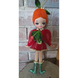 Puede incluir: Adorno de muñeca vintage con cabello de hilo naranja, detalles de hojas verdes y un vestido rojo. La muñeca tiene una cara pintada, piernas de madera y botas con purpurina verde. Se adjunta una cuerda dorada para colgar.