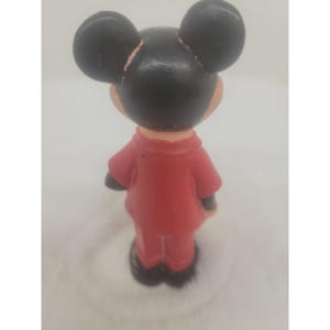 May include: A red and black rubber figurine of Mickey Mouse wearing a red jacket and pants. The figurine is standing with its back to the camera.