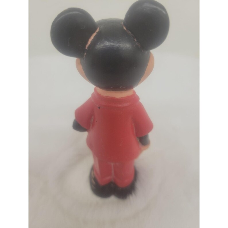 May include: A red and black rubber figurine of Mickey Mouse wearing a red jacket and pants. The figurine is standing with its back to the camera.